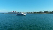A Large White And Blue Yacht Sailing Along The Vast Blue Ocean Water In Front Of The Queen Mary Ship Surrounded By Lush Green Trees, Buildings And Cranes At Shoreline Park In Long Beach California 