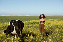 Girl With Curly Hair Posing In A Flowers Field Next To A Cow