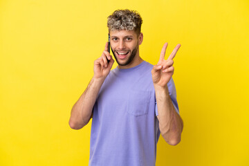 Wall Mural - Young caucasian man using mobile phone isolated on yellow background smiling and showing victory sign