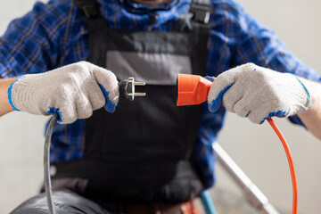 Hands of a man wearing work gloves hold an extension cord and cable connection of two plugs plugging the equipment into electricity.