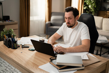 Middle-aged man with beard intently typing text on laptop keyboard. He loves his job, so he is responsible. On the table is cup of coffee, phone, books, calculator and stationery.