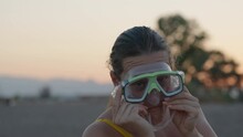 The Girl Tries On A Mask For Scuba Diving Against The Background Of The Sunset Sky, And Makes A Face
