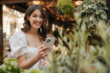 Wall Mural - Smiling young caucasian woman looking at camera, holding phone standing outdoors. Brunette spends her leisure time among plant market. Concept of enjoying moment, technology