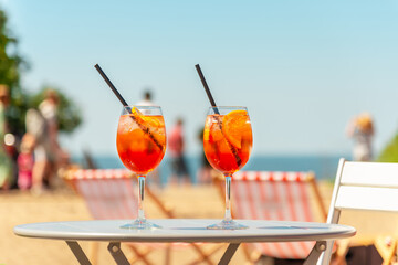 Two glasses of orange spritz aperol drink cocktail on table outdoors with sea and trees view blurred people background.