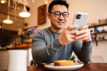 Wall Mural - Adult asian man using cellphone while having breakfast in cafe indoors