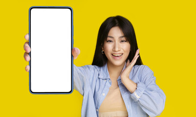 Beautiful asian girl wowing facial expression holding smartphone with white screen happy to introduce new app, game, win, isolated on yellow background. Product placement. Mobile app advertisement