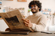 Indian young curly man wearing eyeglasses reading newspaper in cafe