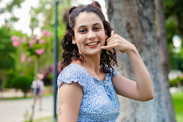 Young brunette girl smiling happy wearing summer dress on city park, outdoors makes a phone gesture and laughs looking at the camera.