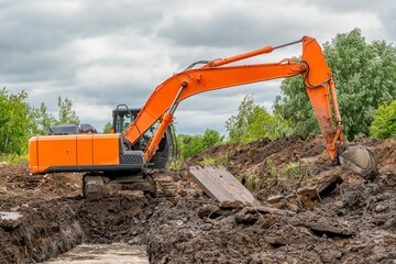 Canvas Print - Orange excavator digs trench in ground against backdrop of green trees and blue summer sky.