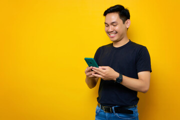 Wall Mural - Smiling young Asian man in casual t-shirt using mobile phone and looking at gadget screen isolated on yellow background. People lifestyle concept