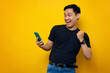 © Bangun Stock Photo - Excited young Asian man in casual t-shirt holding mobile phone, making winner gesture, getting good news isolated on yellow background. People lifestyle concept