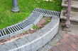 iron drainage grate on the roadside with green grass and stone pebbles at the granite curb along the pedestrian pavement of stone tiles near stairs with steps on city park, nobody.