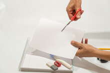 Hand Cutting With Scissors A Sheet Of Paper, Eraser And Liquid Paper On The Table