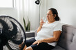 © kleberpicui - Elderly woman sitting on sofa in living room cooling off with floor fan trying to relieve heat of hot summer weather. Elderly woman drinking a glass of water on a hot day.