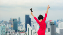 Back Side Of A Short-haired Business Women Wearing A Red Dress, Holding A Tablet, Jumping Joyfully, Dancing On A Rooftop With Blur Background Of High-rise Buildings.