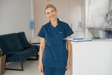 Sticker - Smiling doctor with clipboard standing near reception in clinic hall and looking camera