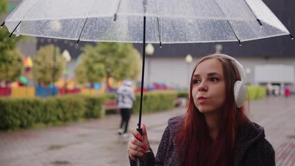 Wall Mural - Young beautiful woman with transparent umbrella dancing in rain on city street. Pretty female in wireless headphones listening to music, sheltering with umbrella from rain on walk in city park.