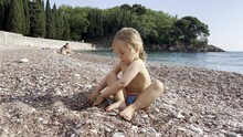Little Girl Digs A Hole With Her Hands On A Pebble Beach Near The Water