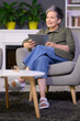 Handsome middle aged woman with grey hair at apartment with tablet in hands. Happy and smiling aged woman using tablet sitting in chair in living room at home. Attractive mid aged woman using internet