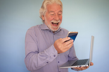 Wall Mural - Cheerful bearded senior man holding laptop while using mobile phone.  70 years old man laughing in video chat enjoying technology and social