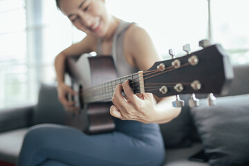 Beautiful asian woman smile while playing acoustic guitar, sitting on sofa at home.