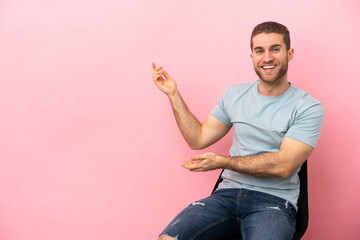 Poster - Young man sitting on a chair over isolated pink background extending hands to the side for inviting to come