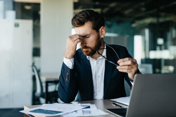 Exhausted businessman sitting in front of laptop at modern office, holding glasses and rubbing nose bridge