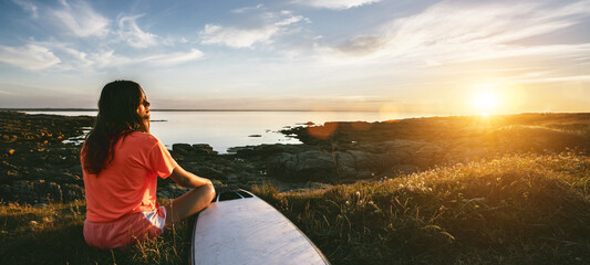 young surfer girl sitting at the ocean coast near her surfboard, watching the sunset and enjoy the nature while deep breathing