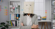 Young Asian businesswoman sit on desk wear draw sad mask paper craft bag feel bad mood with fail work project at office. Anonymous young lady mad and confuse with overwork, Work mental health.