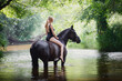 portrait of beautiful young blond woman sitting on black horse in river in summer