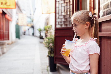 Little caucasian girl in trendy summer clothes. Carefree kid relaxing outdoor in Chinatown at hot summer day. Positive model holding and drinking fresh cocktail smoothie drink in plastic cup with