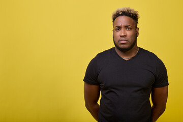 Wall Mural - Calm black guy in a black T-shirt looks at the camera. An African-American man with a serious expression put his hands behind his back on a yellow background