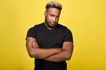 Wall Mural - Strong guy with big muscles stands in a T-shirt on a yellow background and is sad. A disgruntled serious black man stands in a closed position, with his hands folded on his chest cross to cross. 