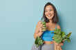 © wayhome.studio  - Indoor shot of cheerful dark haired Asian woman holds freshly made smoothie from vegetables carries net bag wears cropped top and leggings isolated over blue background blank space for promo