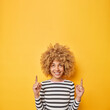 © wayhome.studio - Smiling happy woman with curly blonde hair points fingers up shows advertisement on top wears casual striped jumper isolated over yellow background suggests to go upwards. Big news announcement