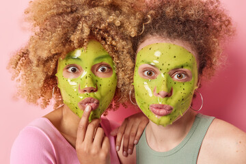 Wall Mural - Portrait of surprised curly haired young women keep lips folded undergo beauty procedures apply green sheet mask on face isolated over pink background. Wellness and cosmetic treatments concept