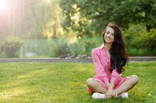 The Girl In A Pink Dress Sits On The Grass With Her Legs Crossed