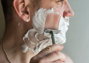 A handsome man shaves with a T-shaped metal chrome razor in natural light