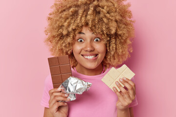 Wall Mural - Positive curly haired woman holds two bars of sweet chocolate has sugar addiction bites lips looks with happy surprised expression isolated over pink background. Female suggests appetizing snack
