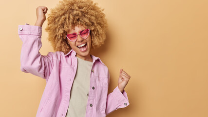 Wall Mural - Joyful happy woman with curly hair dances carefree enjoys moment shakes arms wears trendy heart shaped sunglasses and jacket isolated over brown background with blank space for your advertisement