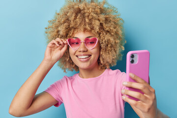 Wall Mural - Good looking cheerful woman wears pink heart shaped sunglasses and casual t shirt smiles gladfully makes video call makes photos isolated over blue background. People joy happiness technology