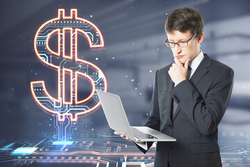 Poster - Attractive thoughtful young european businessman with laptop looking at creative glowing dollar sign hologram on blurry office interior background. Online banking, technology and currency concept.