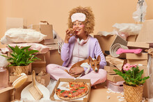 Positive Curly Haired Beautiful Woman Dressed In Domestic Clothes Eats Delicious Pizza Poses With Favorite Dog Relocates In New Dwelling Surrounded By Cardboard Boxes On Floor With Possession.