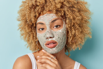 Wall Mural - Close up portrait of curly woman touches jawline applies nourishing clay mask to reduce blackheads looks directly at camera takes care of facial skin isolated over blue background. Facial treatments