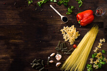 Various Sorts Of Pasta On Top Of A Dark Wooden Background. Farfalle, Fusilli, Spaghetti, Bell Pepper, Corn Salad, Garlic Cloves, Red, Green Or Yellow Pepper.
