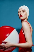 Delighted Woman In A Bathing Cap Red Swimsuit With A Striped Ball Turns Back Posing On A Blue Background In The Studio. Summer Holiday Concept