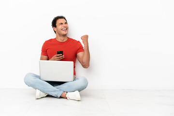 Wall Mural - Caucasian handsome man with a laptop sitting on the floor with phone in victory position