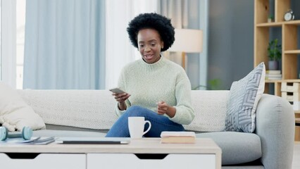 Wall Mural - Happy African American woman texting on a phone while relaxing on a sofa and drinking coffee at home. Smiling black woman browsing social media and laughing, planning on how to spend her free time