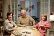 © pressmaster - Three senior happy people in shirts looking at you while sitting by table with boards and other supplies for leisure games in living room