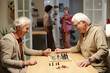 © pressmaster - Two cheerful retired men in casualwear looking at chessboard with black and white checkeres and figures before start playing game